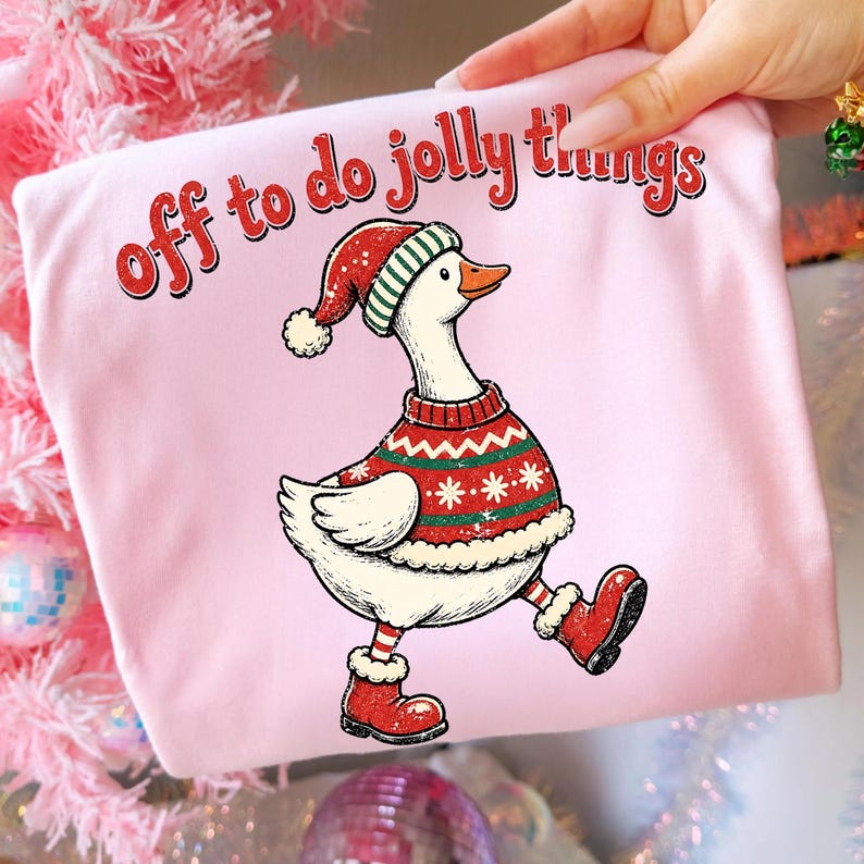 May include: A light pink t-shirt featuring a graphic of a white goose wearing a red and green patterned sweater, a matching Santa hat, and red boots. The text above the goose reads "off to do jolly things" in a red, distressed font style.