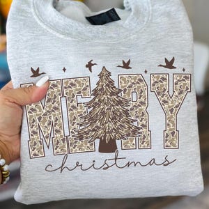 May include: A light gray sweatshirt with the words "MERRY christmas" in a brown camouflage pattern. A brown Christmas tree is in the center of the word "MERRY", with brown birds flying above the letters.