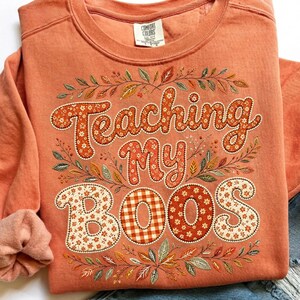 Pu&ograve; includere: Una felpa girocollo color corallo con la scritta Teaching My BOOS in un carattere grassetto e arrotondato. Le lettere sono riempite con motivi floreali bianchi e quadretti vichy arancioni, circondate da un bordo di foglie autunnali.