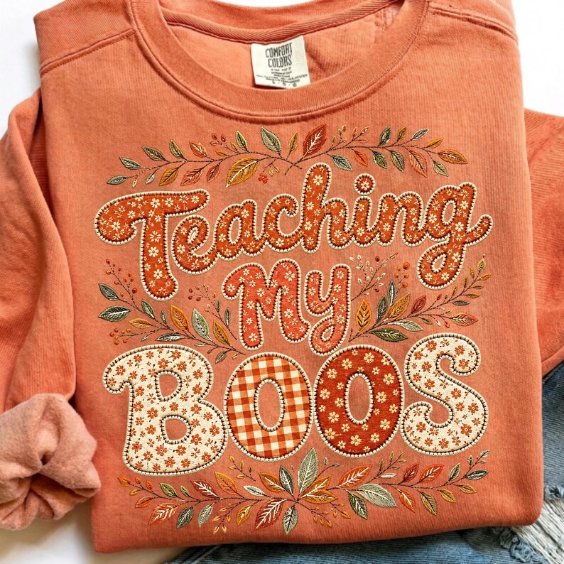 Pu&ograve; includere: Una felpa girocollo color corallo con la scritta Teaching My BOOS in un carattere grassetto e arrotondato. Le lettere sono riempite con motivi floreali bianchi e quadretti vichy arancioni, circondate da un bordo di foglie autunnali.