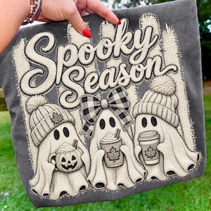May include: A dark gray fabric featuring an embroidered design of three ghosts. The text Spooky Season is at the top. The ghosts wear knit hats and a plaid bow, holding a pumpkin mug and coffee cups with maple leaf designs on a fence background.