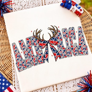 May include: A white t-shirt featuring the word AMERICA in a red, white, and blue camouflage pattern. The letter M is replaced by a pair of brown deer antlers and a matching camouflage bow. Small red four-pointed star accents surround the text.