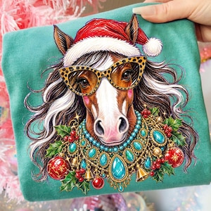 May include: A teal t-shirt featuring a graphic illustration of a brown horse wearing a red and white Santa hat, leopard print cat-eye glasses, and an elaborate turquoise and gold statement necklace adorned with red Christmas ornaments and holly.