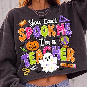 May include: A charcoal gray long-sleeve sweatshirt featuring a colorful graphic with the text "You Can't SPOOK ME I'm a TEACHER". The design includes a pumpkin, a globe, scissors, a pencil, an eraser, a ruler, and a ghost with small flower patterns.