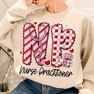 May include: A beige sweatshirt with a graphic design featuring the letters "NP" in red and white, with a stethoscope, hearts, and the words "Nurse Practitioner" in black script. The design has a crochet-like texture.