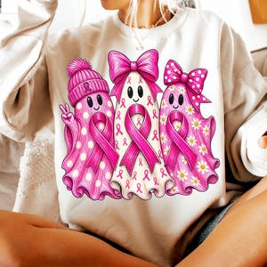 Puede incluir: Una sudadera beige de cuello redondo con un gr&aacute;fico de tres fantasmas decorados con lazos rosas de concienciaci&oacute;n sobre el c&aacute;ncer de mama. El fantasma de la izquierda lleva un gorro rosa con pomp&oacute;n, el central un lazo rosa grande y el de la derecha un lazo rosa.