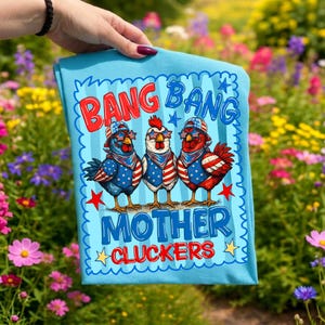 May include: A light blue t-shirt featuring a graphic of three chickens wearing patriotic star-spangled hats, bandanas, and sunglasses. The text reads BANG BANG MOTHER CLUCKERS in red and blue lettering, surrounded by a scalloped border and stars.