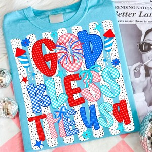 K&ouml;nnte beinhalten: Ein hellblaues T-Shirt mit einem Grafikdruck und dem Text GOD BLESS THE USA. Das Design umfasst rote, wei&szlig;e und blaue Muster, Punkte, Karos, Feuerwerk, Sterne und Raketenillustrationen auf einem gestreiften Hintergrund.