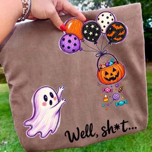 May include: A brown tote bag featuring a graphic of a white ghost reaching for a pumpkin bucket filled with candy, held by a cluster of colorful balloons with polka dot and star patterns. The text "Well, sh*t..." is printed in black script.