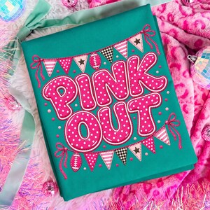 K&ouml;nnte beinhalten: Ein t&uuml;rkisfarbenes Stoffquadrat mit dem Text PINK OUT in gro&szlig;en, pinkfarbenen Buchstaben mit wei&szlig;en Punkten. Das Design enth&auml;lt zwei Wimpelketten mit pinken, wei&szlig;en und schwarzen Mustern, durchsetzt mit zwei kleinen wei&szlig;en Football-Grafiken.