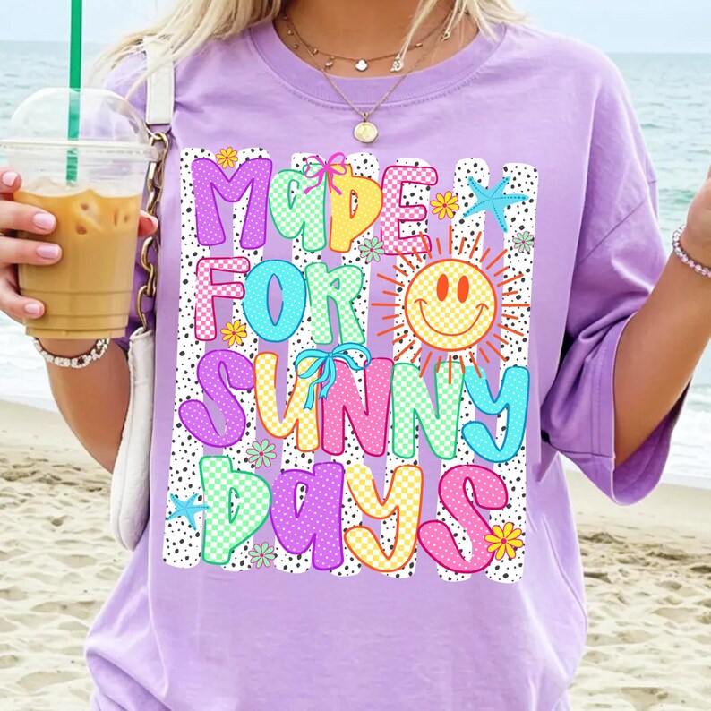 May include: A light purple t-shirt featuring a graphic print with the text "MADE FOR SUNNY DAYS" in colorful, patterned bubble letters. The design includes a yellow smiley face sun, small flowers, blue starfish, and pink bows over black stripes.