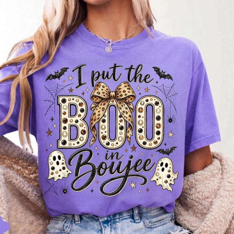 Pu&ograve; includere: Una t-shirt a maniche corte color lavanda con una stampa grafica che riporta il testo I put the BOO in Boujee. Il design include un fiocco con stampa leopardata, due fantasmi, ragnatele, pipistrelli e piccole stelle nei toni nero, oro e bianco.