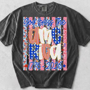 K&ouml;nnte beinhalten: Ein anthrazitfarbenes Kurzarm-T-Shirt mit einem Grafikdruck und dem Text God Bless The America 1776-2026. Das Design zeigt vertikale Streifen mit Blumen- und Punktemustern in Rot, Wei&szlig; und Blau sowie Sternmotive.