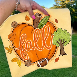 Puede incluir: Una camiseta de color amarillo p&aacute;lido con un estampado gr&aacute;fico de una gran calabaza naranja con la palabra fall escrita en letra cursiva blanca. El dise&ntilde;o incluye un &aacute;rbol verde, un bal&oacute;n de f&uacute;tbol americano, nueces y hojas dispersas.