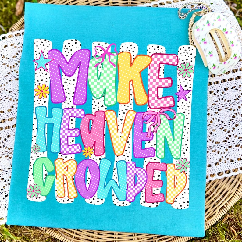 May include: A bright turquoise t-shirt featuring the text "MAKE HEAVEN CROWDED" in colorful, patterned block letters. The design includes white vertical stripes with black dots, small pink bows, stars, and flowers surrounding the lettering.