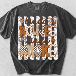 Pode incluir: Uma t-shirt de manga curta cinzento-carv&atilde;o com uma impress&atilde;o gr&aacute;fica com o texto Kinda BOUJEE Kinda BROKE. O design inclui riscas verticais brancas com pontos pretos, padr&otilde;es axadrezados castanhos e beges, pequenas flores, estrelas e la&ccedil;os.