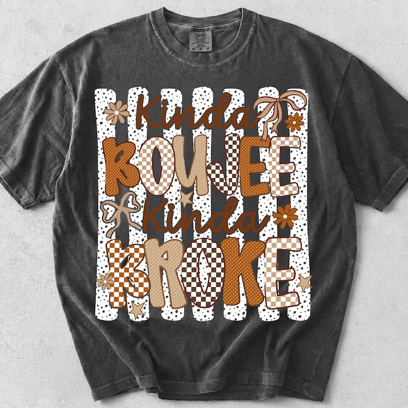 Pode incluir: Uma t-shirt de manga curta cinzento-carv&atilde;o com uma impress&atilde;o gr&aacute;fica com o texto Kinda BOUJEE Kinda BROKE. O design inclui riscas verticais brancas com pontos pretos, padr&otilde;es axadrezados castanhos e beges, pequenas flores, estrelas e la&ccedil;os.