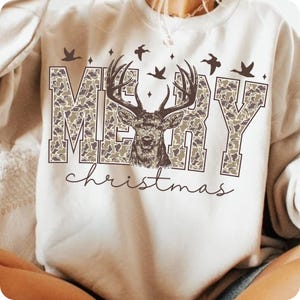 May include: Cream-colored sweatshirt with a camouflage-patterned "MERRY" design and a deer graphic. The word "christmas" is written in a cursive font below the main design. Includes flying birds and stars.