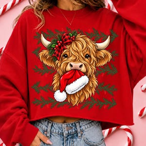 May include: A bright red long-sleeve sweatshirt featuring a graphic of a shaggy Highland cow with a red and black buffalo plaid bow on its head and a red Santa hat held in its mouth, set against a background pattern of green pine branches.
