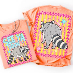 Op de afbeelding: Een perzikkleurig t-shirt met een afbeelding van een wasbeer en de tekst SEE YA SOON in lichtblauwe letters. Het ontwerp bevat een geel en oranje gestreepte achtergrond met roze geschulpte randen en kleine lichtblauwe en gele sterren.
