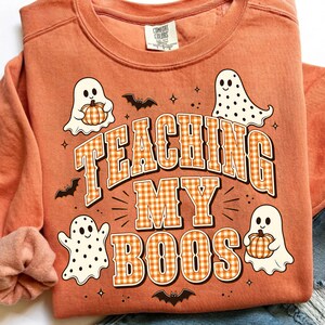 May include: An orange crewneck sweatshirt featuring a graphic print with the text TEACHING MY BOOS in orange and white gingham letters. The design includes three white ghosts, two holding orange pumpkins, small black bats, and tiny stars.