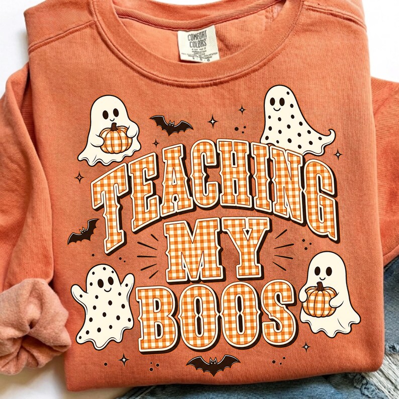 May include: An orange crewneck sweatshirt featuring a graphic print with the text TEACHING MY BOOS in orange and white gingham letters. The design includes three white ghosts, two holding orange pumpkins, small black bats, and tiny stars.