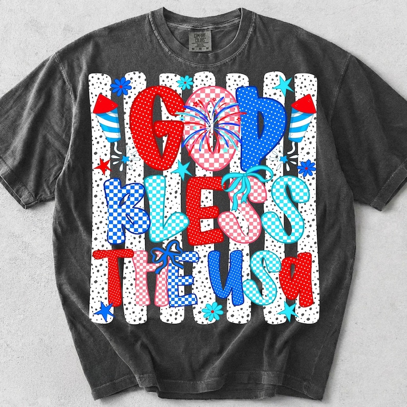 K&ouml;nnte beinhalten: Ein anthrazitfarbenes T-Shirt mit einem Grafikdruck und dem Text GOD BLESS THE USA. Das Design zeigt rote, wei&szlig;e und blaue Muster, Punkte, Karos, Feuerwerk, Sterne, Blumen und Raketensymbole auf vertikalen Streifen.