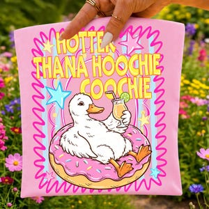 May include: A light pink fabric item featuring a graphic of a white duck floating on a pink donut with sprinkles. The duck holds a cocktail with a lime slice. The text reads HOTTER THANA HOOCHIE COOCHIE in yellow letters with a pink zigzag border.