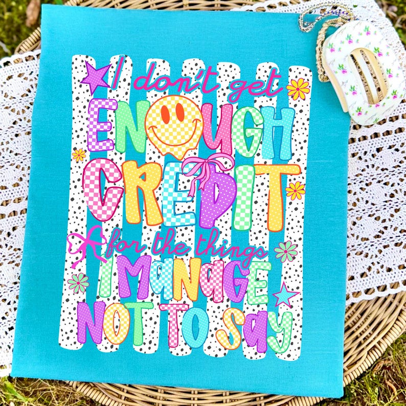Pode incluir: Uma t-shirt turquesa brilhante com uma estampa gr&aacute;fica com o texto I don't get ENOUGH CREDIT for the things I MANAGE NOT TO SAY. O design inclui um rosto sorridente xadrez amarelo, la&ccedil;os cor-de-rosa, estrelas e flores sobre listras.