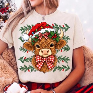 Puede incluir: Una camiseta de color crema con un gr&aacute;fico de una vaca Highland marr&oacute;n que lleva un gorro de Pap&aacute; Noel rojo y un lazo a cuadros rojos y blancos. La vaca est&aacute; rodeada de ramas de pino verdes, bayas rojas y bombillas navide&ntilde;as de colores.