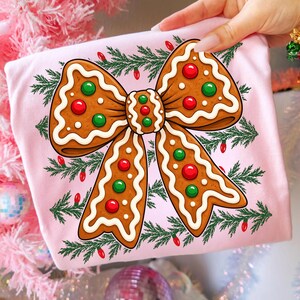 Op de afbeelding: Een lichtroze T-shirt met een grote peperkoek in de vorm van een strik, versierd met wit glazuur en rode en groene snoepjes. Het ontwerp is omringd door groene dennentakken en kleine rode kerstlichtjes op de stof.