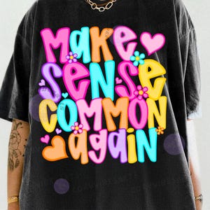May include: A black t-shirt with the phrase "Make Sense Common Again" in colorful bubble letters. The text includes pink, orange, blue, yellow, and purple colors, with heart and flower accents. The shirt has a vintage, worn look.