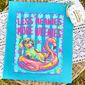 May include: A turquoise t-shirt featuring a graphic of a dachshund wearing heart-shaped sunglasses while floating on a pink flamingo pool float. The text reads LESS MEANIES MORE WEENIES in pink letters, surrounded by small yellow and pink stars.