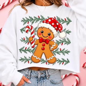 Puede incluir: Una sudadera corta blanca con un gr&aacute;fico de un hombre de jengibre que lleva un gorro de Pap&aacute; Noel rojo con lunares blancos y una pajarita roja. El hombre de jengibre sostiene un bast&oacute;n de caramelo a rayas rojas y blancas sobre ramas de pino.