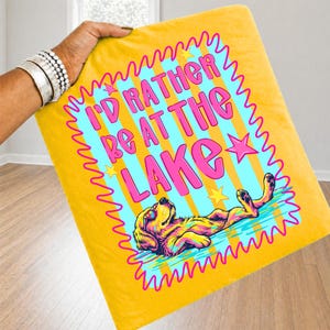 May include: A yellow throw blanket featuring a graphic of a dog floating in water with the text I'D RATHER BE AT THE LAKE in pink letters. The design includes a light blue background with yellow stripes, pink star accents, and a wavy pink border.