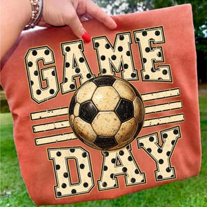 May include: A burnt orange t-shirt featuring a graphic print with the text GAME DAY in large, cream-colored block letters with black polka dots. A central soccer ball graphic with a distressed texture sits between the words GAME and DAY.