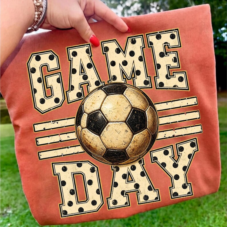 May include: A burnt orange t-shirt featuring a graphic print with the text GAME DAY in large, cream-colored block letters with black polka dots. A central soccer ball graphic with a distressed texture sits between the words GAME and DAY.