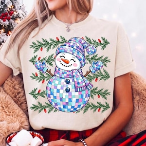 May include: A cream-colored t-shirt featuring a graphic of a snowman made of iridescent disco ball tiles. The snowman wears a matching hat, scarf, and mittens, set against a background of green pine branches and small red Christmas light bulbs.