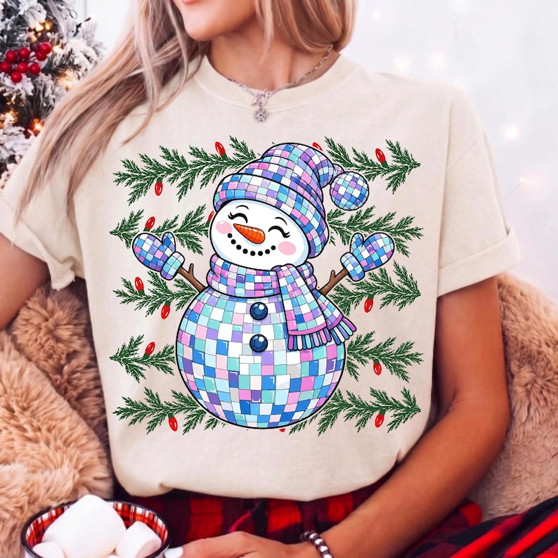 May include: A cream-colored t-shirt featuring a graphic of a snowman made of iridescent disco ball tiles. The snowman wears a matching hat, scarf, and mittens, set against a background of green pine branches and small red Christmas light bulbs.