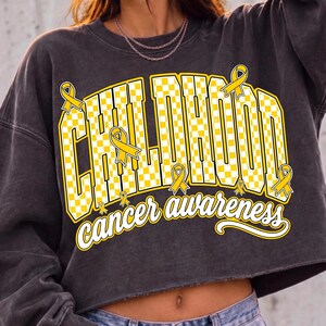Puede incluir: Una sudadera corta de color gris carb&oacute;n con un estampado gr&aacute;fico que muestra el texto CHILDHOOD en un patr&oacute;n de cuadros amarillos y blancos, acompa&ntilde;ado de las palabras cancer awareness en letra blanca y varios lazos amarillos.