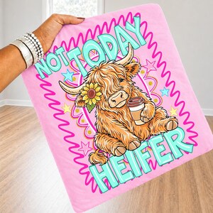 May include: Pink blanket with a cartoon Highland cow design. The cow has orange fur, a sunflower, and holds a coffee cup. The blanket has the text "NOT TODAY HEIFER" in turquoise and pink.