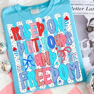 May include: A light blue t-shirt featuring a graphic print with the text "Raised on Dirt Roads and Freedom" in red, blue, and pink checkered and polka-dot patterns. The design includes firework illustrations, stars, and bows on a striped background.