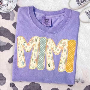 May include: A purple short-sleeve t-shirt with a crew neckline and a Comfort Colors brand label. The front features the text MIMI in large letters, with each letter filled with a different pattern of small flowers, white stars on sage, or white stars on yellow.