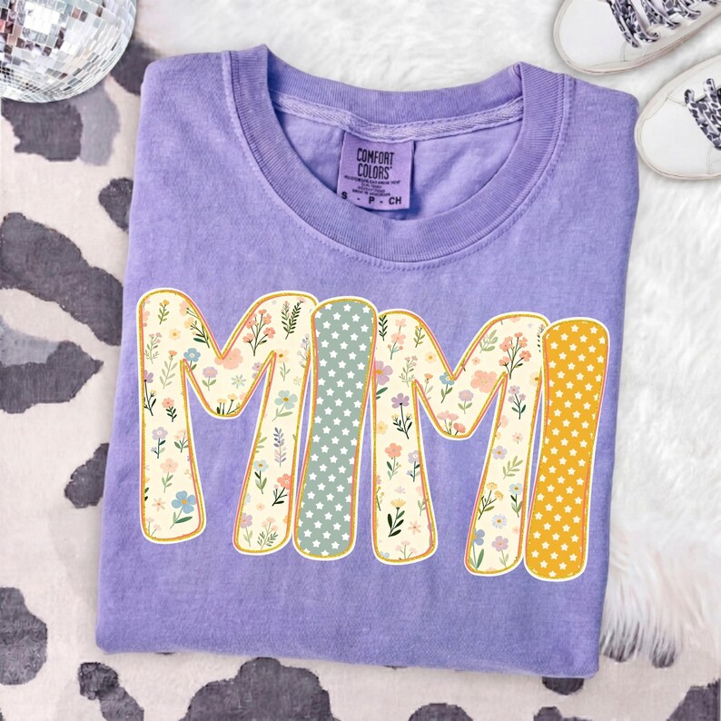 May include: A purple short-sleeve t-shirt with a crew neckline and a Comfort Colors brand label. The front features the text MIMI in large letters, with each letter filled with a different pattern of small flowers, white stars on sage, or white stars on yellow.