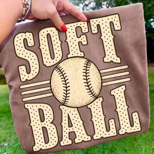 以下が含まれることがあります： 大きなクリーム色の水玉模様のブロック体で「SOFT BALL」という文字がプリントされた茶色のTシャツ。縫い目が見える野球ボールのグラフィックが中央に配置され、その両側に3本の水平線が描かれています。