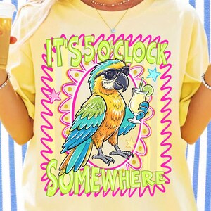 May include: Yellow t-shirt with a colorful parrot graphic wearing sunglasses and holding a cocktail. The text "IT'S 5 O'CLOCK SOMEWHERE" is printed above and below the parrot in green and pink. The design is surrounded by a pink wavy border.