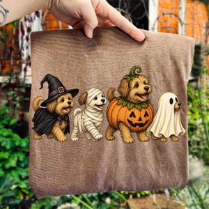 Puede incluir: Una camiseta marr&oacute;n con un gr&aacute;fico de cuatro perros con disfraces de Halloween: uno con sombrero de bruja y capa negra, otro envuelto en vendas de momia blancas, otro dentro de una calabaza naranja tallada y otro cubierto por una s&aacute;bana de fantasma blanca.