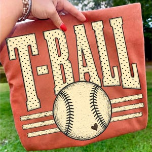Pode incluir: Uma t-shirt cor de laranja queimado com o texto T-BALL em letras grandes de cor creme com um padr&atilde;o de pontos pretos. Abaixo do texto, h&aacute; um gr&aacute;fico de uma bola de basebol com costuras vis&iacute;veis e um pequeno cora&ccedil;&atilde;o preto.