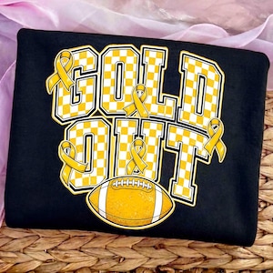 Puede incluir: Una camiseta negra con el texto GOLD OUT en letras grandes de bloque con un patr&oacute;n de cuadros amarillos y blancos. El dise&ntilde;o incluye varios lazos amarillos de concienciaci&oacute;n integrados en las letras y un gr&aacute;fico de un bal&oacute;n de f&uacute;tbol.