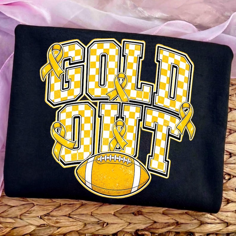 Puede incluir: Una camiseta negra con el texto GOLD OUT en letras grandes de bloque con un patr&oacute;n de cuadros amarillos y blancos. El dise&ntilde;o incluye varios lazos amarillos de concienciaci&oacute;n integrados en las letras y un gr&aacute;fico de un bal&oacute;n de f&uacute;tbol.